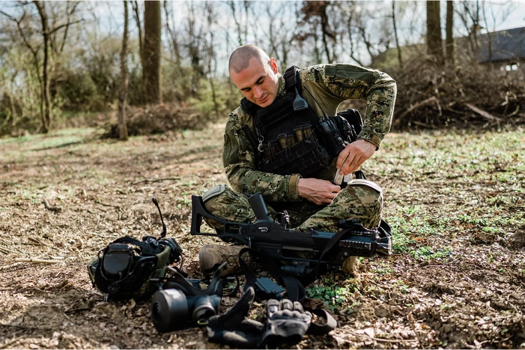 A soldier preparing his ballistic clothing, adjusting his bulletproof vest while sitting on the ground in a forest setting