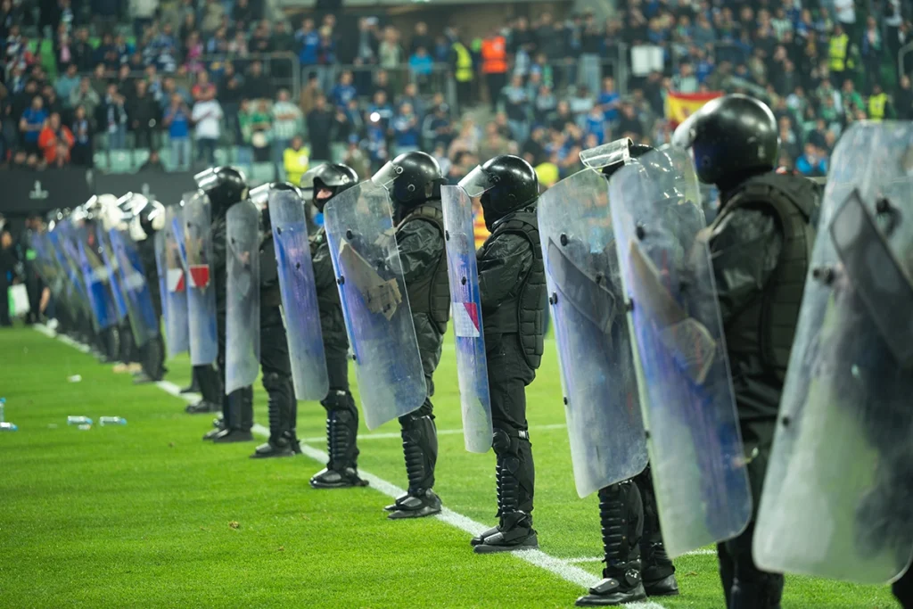 A line of riot police on a stadium pitch, where the basic protective equipment is a riot shield.
