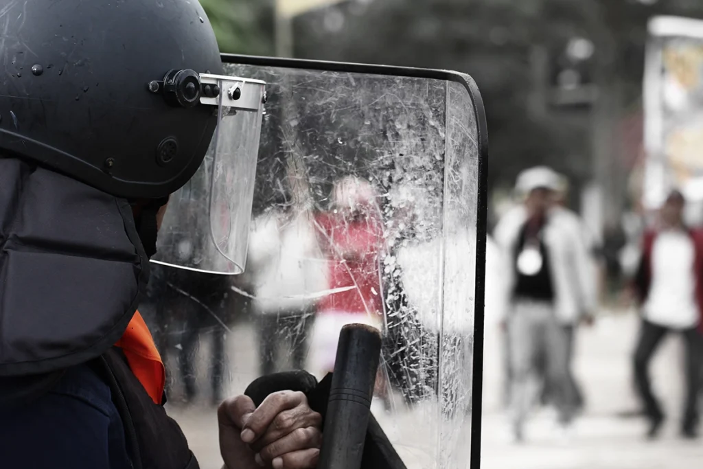 A riot police officer in a helmet holding a baton and a polycarbonate riot shield during street clashes.